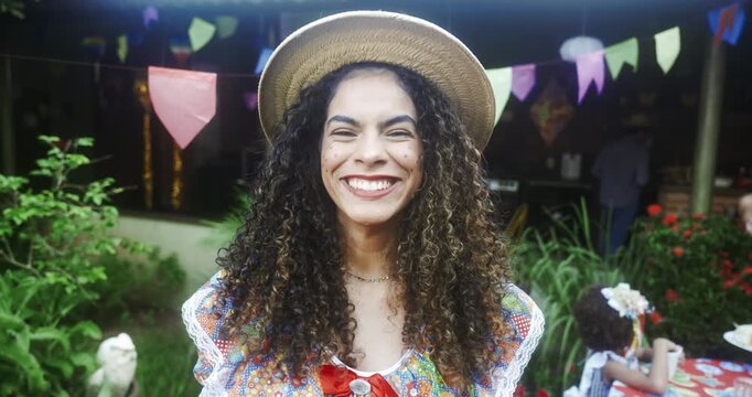 A cheerful Brazilian woman with curly hair and a straw hat smiles while celebrating Festa Junina at a traditional outdoor party decorated with colorful flags and festive ornaments
