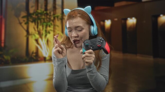 Woman with headphones and cat ear headband holds a gamepad and makes crossed fingers gesture in a building lobby, smiling toward camera; playful anticipation.