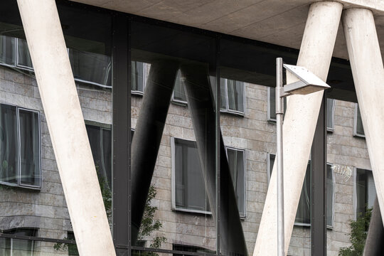 angled concrete columns shape exterior architecture beneath an overhead plane, where urban shadow and geometric structure press against the facade.