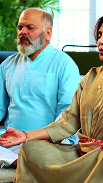 Indian senior couple doing omkar pranayama or yoga at home in the morning sitting with folded legs in modern living room chanting aum, practicing mindful breathing exercise for health