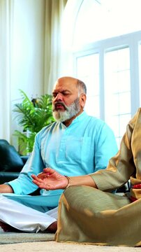 Indian senior couple doing omkar pranayama or yoga at home in the morning sitting with folded legs in modern living room chanting aum, practicing mindful breathing exercise for health