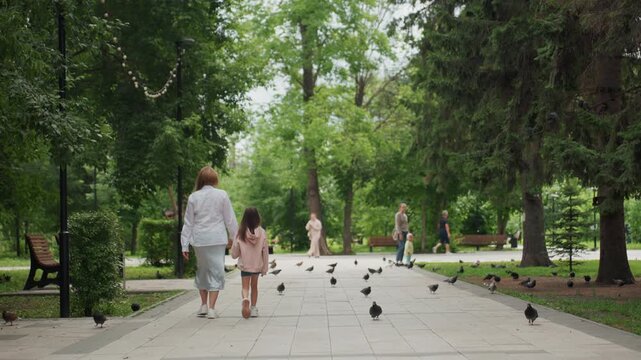 Mom and daughter walking through park, pigeons scattering across paved walkway, green trees and benches lining path, summer light, holding hands, warm tender bond, casual clothing, serene family