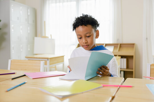 A young boy attentively reads a workbook in a bright classroom with colorful stationery scattered on desks. Back to school and education concept