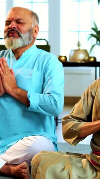 Indian senior couple doing omkar pranayama or chanting mantra or yoga at home in the morning sitting with folded legs in modern living room, practicing mindful breathing for health
