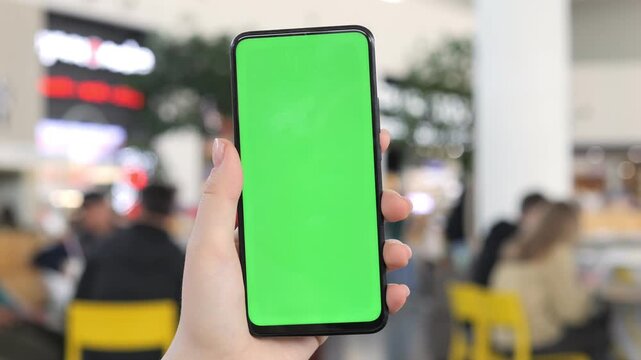 Hand holds phone with green screen. Background shows blurred indoor space with people and chairs. Three keyframes show same scene with slight movement. No animals or plants visible