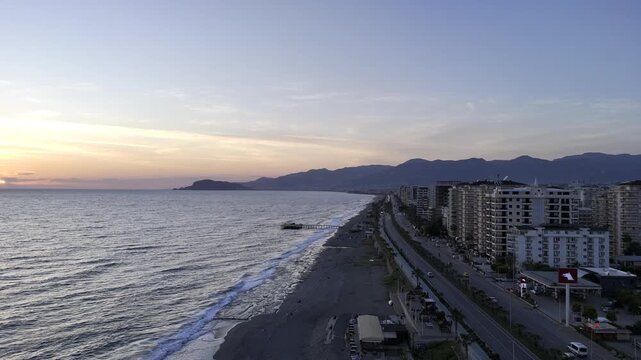 Aerial drone panorama of Mahmutlar, Alanya, at sunset. The footage captures the Mediterranean coastline, sandy beach, coastal highway, and dense urban development. Warm evening light illuminates the h