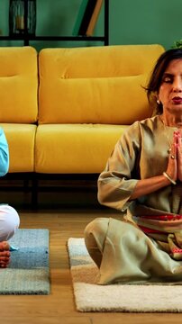 Indian senior couple doing omkar pranayama or chanting mantra or yoga at home in the morning sitting with folded legs in modern living room, practicing mindful breathing for health