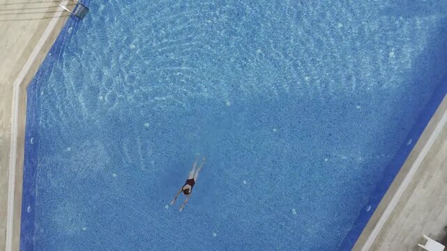 Drone top-down view of a young woman swimming in a crystal clear blue pool at a modern residential complex in Mahmutlar. Sunlit summer day scene with luxury amenities, loungers, umbrellas, and green l