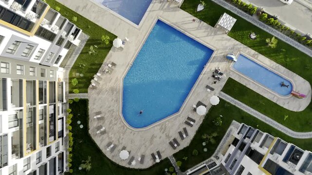 Drone top-down view of a young woman swimming in a crystal clear blue pool at a modern residential complex in Mahmutlar. Sunlit summer day scene with luxury amenities, loungers, umbrellas, and green l