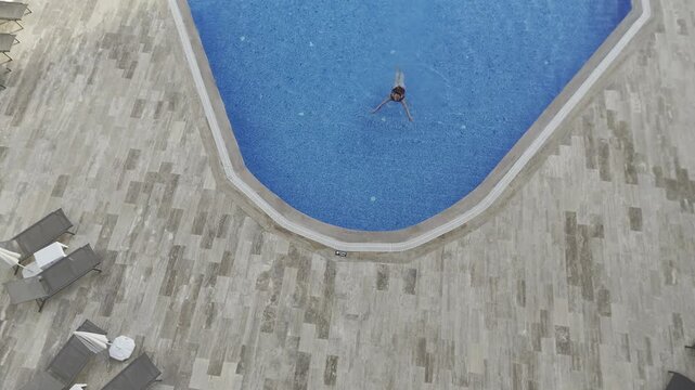 Drone top-down view of a young woman swimming in a crystal clear blue pool at a modern residential complex in Mahmutlar. Sunlit summer day scene with luxury amenities, loungers, umbrellas, and green l