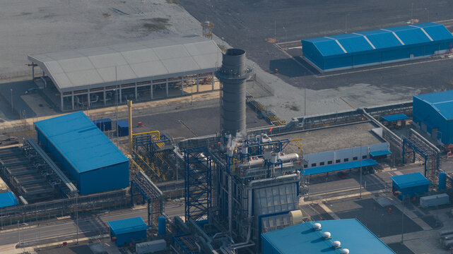 Aerial view of the Gulf Pluak Daeng Power Plant GPD with a tall exhaust stack, blue warehouses, and intricate pipeline systems in Map Yang Phon, Rayong, Thailand.