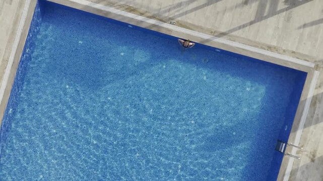 Drone top-down view of a young woman swimming in a crystal clear blue pool at a modern residential complex in Mahmutlar. Sunlit summer day scene with luxury amenities, loungers, umbrellas, and green l