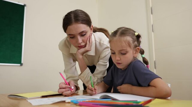 A young female teacher attentively helps a child with schoolwork as they write with colored pencils at a desk in a classroom.