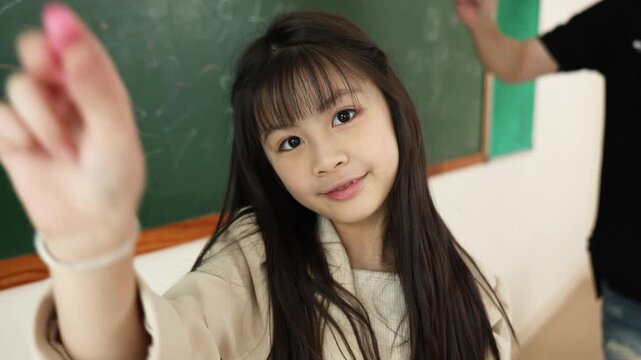 A young student writes math equations on a green chalkboard in a classroom setting.