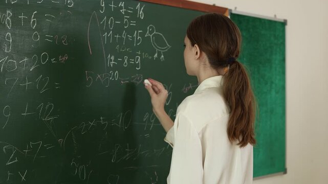 A student solving math problems on a green chalkboard in a classroom, with simple arithmetic equations and a small bird drawing visible.