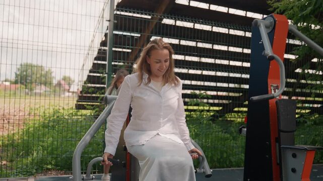 Outdoor park seated mom exercising on machine, white dress, calm expression, urban greenery backdrop, metal fitness equipment, steady breathing and gentle motion showcasing rehabilitation and balance