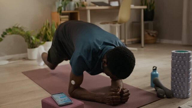 Tracking shot of young adult African American man with diabetes holding plank position during home workout while monitoring glucose readings through mobile app connected to CGM device