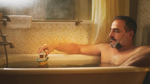 Relaxed mature man playing with toy boat in vintage bathtub with warm lighting