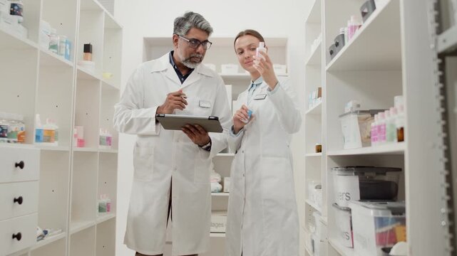 Front view low angle shot of two people wearing white coats, Indian man and Caucasian woman inspecting medications and reading labels while doing stock inventory together in pharmacy storage room