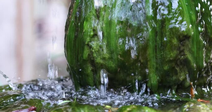 Flowing water splashing over green mossy rock with refreshing droplets and ripples. Natural relaxing scene ideal for spa, wellness, ecology, and background footage