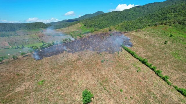 Aerial drone footage of a burning corn crops hill, with many smokes, made to regenerate the lands, in Kwangko area, Dompu regency, Sumbawa island, Indonesia, with mountains and forests behind