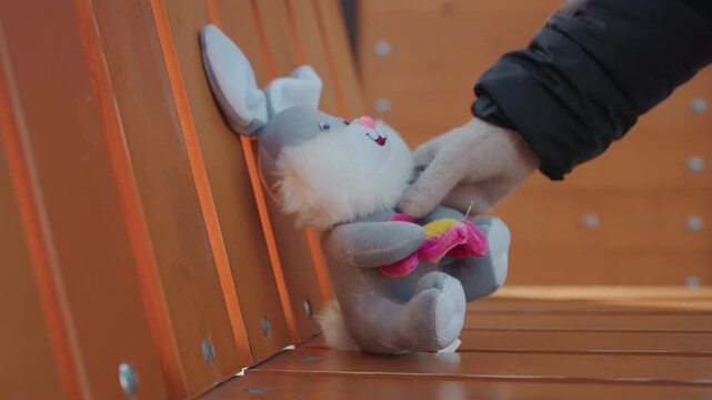 Stuffed bunny on bench, gloved hand removes plush snatcher implied as wintergloved hand takes abandoned toy from wooden park bench near chained swing, deserted playground atmosphere, tension