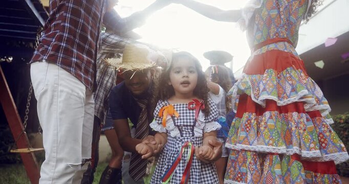 A joyful group of people and children celebrate the traditional Brazilian Festa Junina outdoors, wearing colorful caipira costumes and straw hats while dancing in a circle under bright sunlight