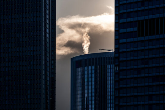 Between dark office buildings, smoke lifts past a glass facade as a tower and urban architecture turn the skyline into a stark pollution scene.