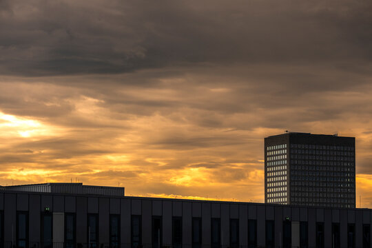 At sunset, clouds gather over the urban skyline while a dark building and tower settle into rooftop silhouette beneath a storm horizon and evening sky.