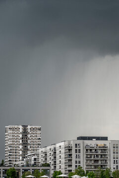 Beyond urban apartment buildings, storm clouds release rain into a gray sky, softening the skyline as windows and facade details hold the weather.