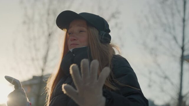 Caucasian woman holds stuffed toy outdoors, examines toy while extending gloved hand, rimlight sunlight on hair, urban buildings in background, cautious stance and alert to possible prowler