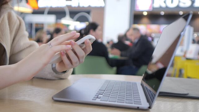 Person uses a smartphone while sitting beside an open laptop. Background shows blurred indoor public space with people. Lighting appears natural and bright. No animals or plants visible
