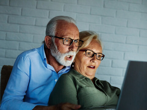 Portrait of an edlerly senior aged couple using laptop at night, watching tv together at home. Shot of a mature couple resting on the couch watching scary movie movie on a laptop, shocked, surpised te