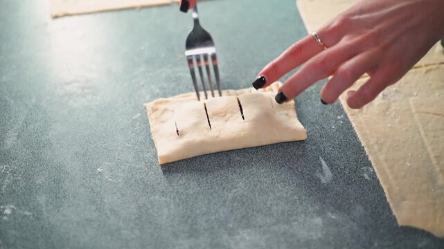 A person with light skin and dark nails uses a fork to create markings on a piece of dough. This action takes place on a clean kitchen counter, highlighting a step in meal preparation