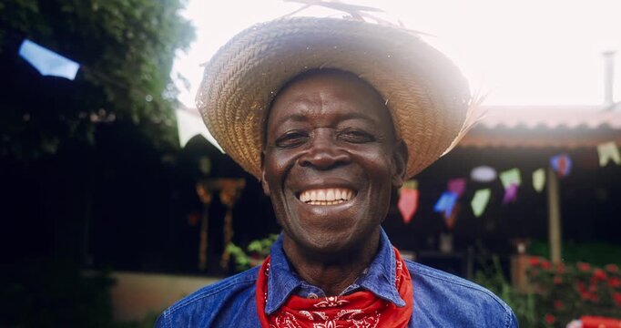 A joyful elderly Black man wearing a traditional straw hat and a red bandana smiles warmly at the camera during a festive outdoor celebration with colorful flags in the background