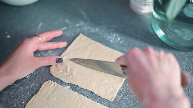 A person with light skin is cutting dough on a countertop in a kitchen. The action focuses on manually shaping the dough pieces while tools and ingredients are visible in the background