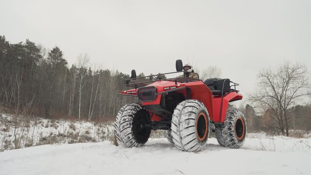 Red atv parked in snowy field, oversized flotation tires with snowpacked tread, frontal angle showing cargo rack and headlamp, rider in helmet and camouflage jacket seated behind controls, engine idle