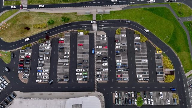 High angle aerial movie of a busy outdoor shopping mall parking lot with rows of parked cars on asphalt spaces, traffic lanes, pedestrian crossing, green grass areas and city road.