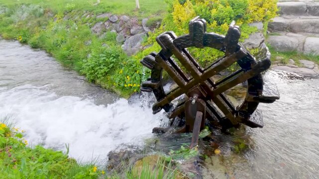 Small wooden water wheel spinning in a clear mountain stream surrounded by green grass and flowers