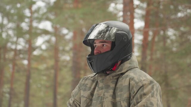 Helmeted rider on atv in forest pauses under pine canopy wearing camo jacket and gloves gripping handlebars with bright LED headlight dusted by snow focused before extreme offroad run