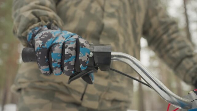 Gloved hand gripping atv handlebar close, blue camo glove on throttle, brake lever and LED bar illuminated, camo jacket and helmeted rider in snowy pine backdrop, tense preparation for extreme ride