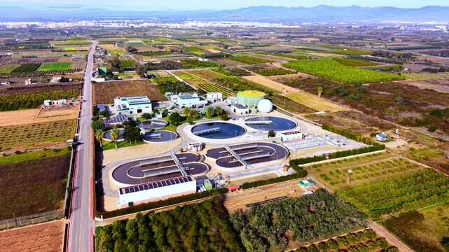 Industrial water purification facility with circular tanks, solar panels, and surrounding agricultural fields under clear sky. Aerial view of modern wastewater treatment plant in rural area.