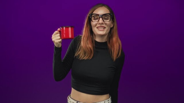 Woman holds red mug up in studio purple backdrop, wearing black turtleneck crop top and glasses; cheerful casual.