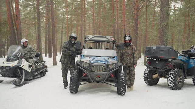 Snow forest atv and snowmobile lineup, three riders in helmets and insulated suits prepare expedition gear, guide checks winch, mechanic inspects suspension, snowflakes falling under tall pine canopy,