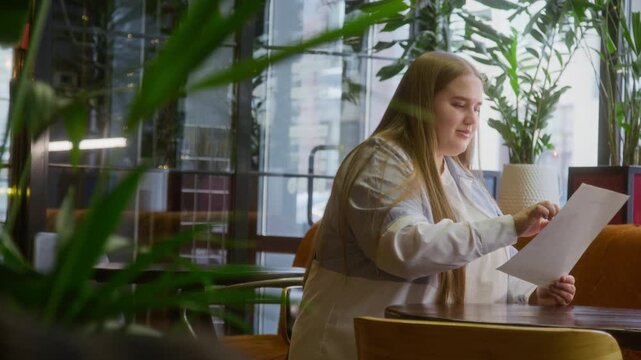 Young woman with long hair sitting at wooden table, carefully studying menu while pointing at it with her finger in cozy, green cafe