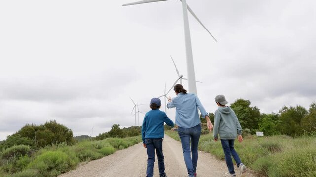 Mother teaching her two sons about renewable energy and sustainability while walking on a country road through a wind farm with large modern turbines generating power