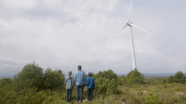Father with sons watching renewable wind turbine energy, learning about sustainability, conservation, and the future of clean electric power generation from nature