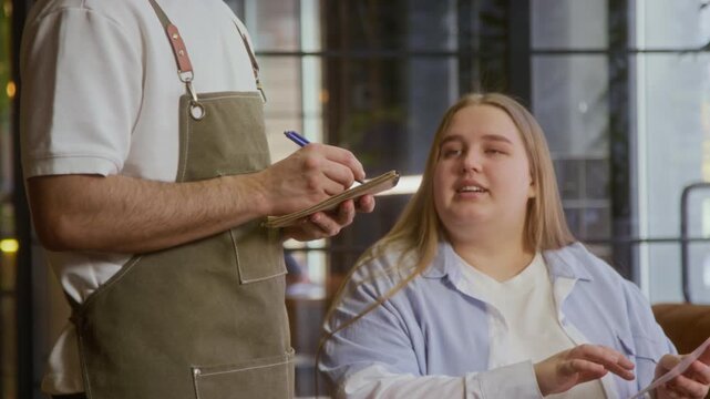 Low angle tilt up shot of young woman telling her order to waiter in apron, taking down notes in notebook at table in modern restaurant