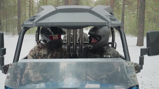 Front view atv duo helmets, two camouflaged riders sit sidebyside inside utv on snowy pine trail, driver grips throttle while passenger scans route, visible radio headset and protective jackets,