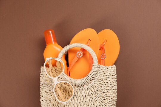 Beach bag with sunglasses, flip flops and bottle of sunscreen on brown background, top view